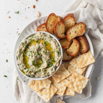 square overhead hero of healthy Mediterranean white bean tuna dip in a bowl on top of a plate with saltines and crostini for serving.