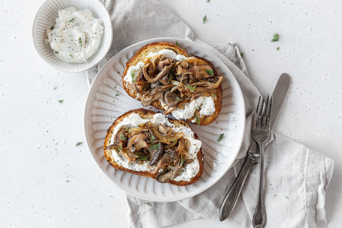 horizontal overhead shot of two wild mushroom tartines on a grey dinner plate with a bowl of spreadable dill goat cheese to the side.