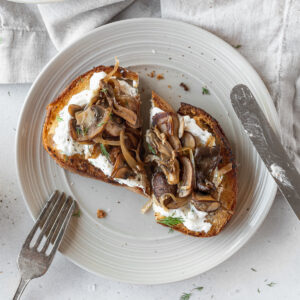 square hero shot of wild mushroom tartine sliced in half on a grey plate with a fork and knife.