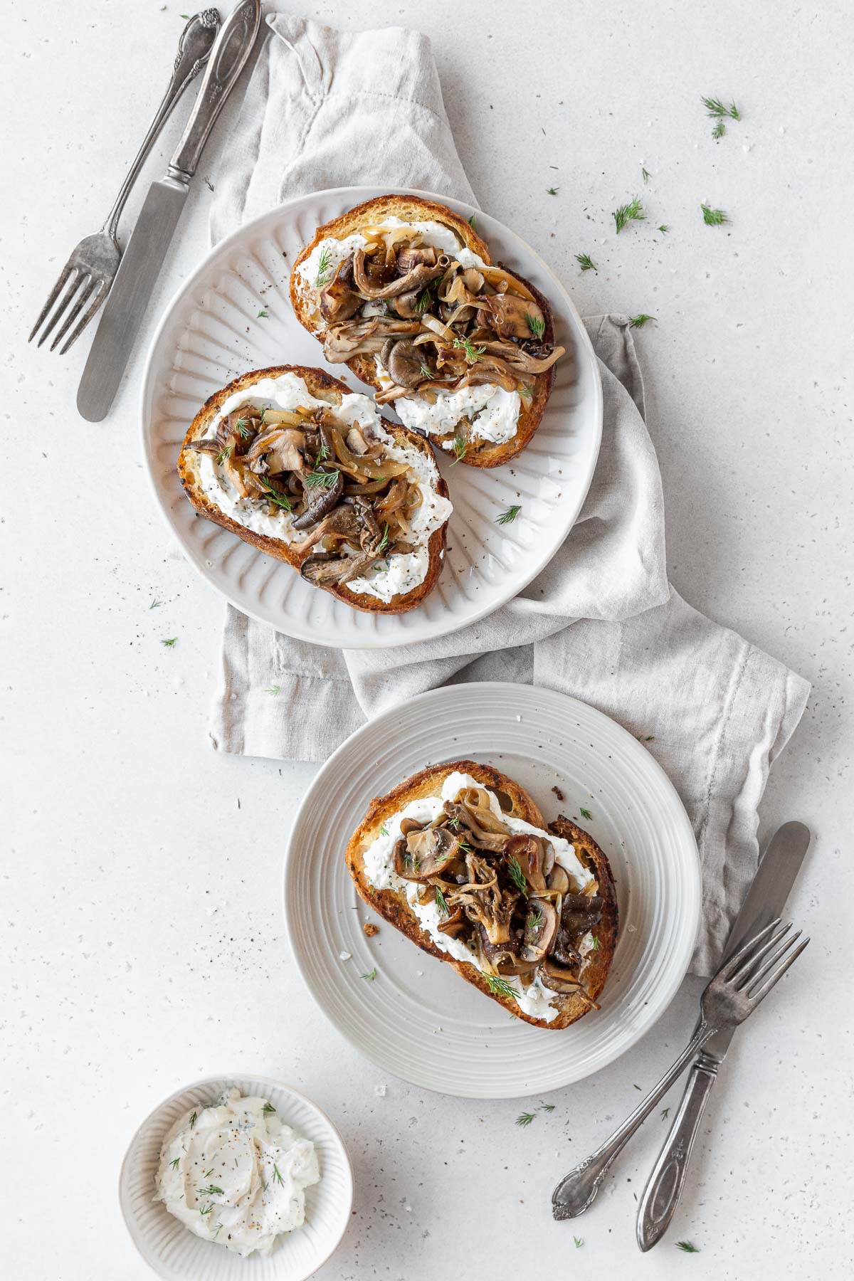 overhead shot of two plates with mushroom tartines on a white table with silver forks and knives.