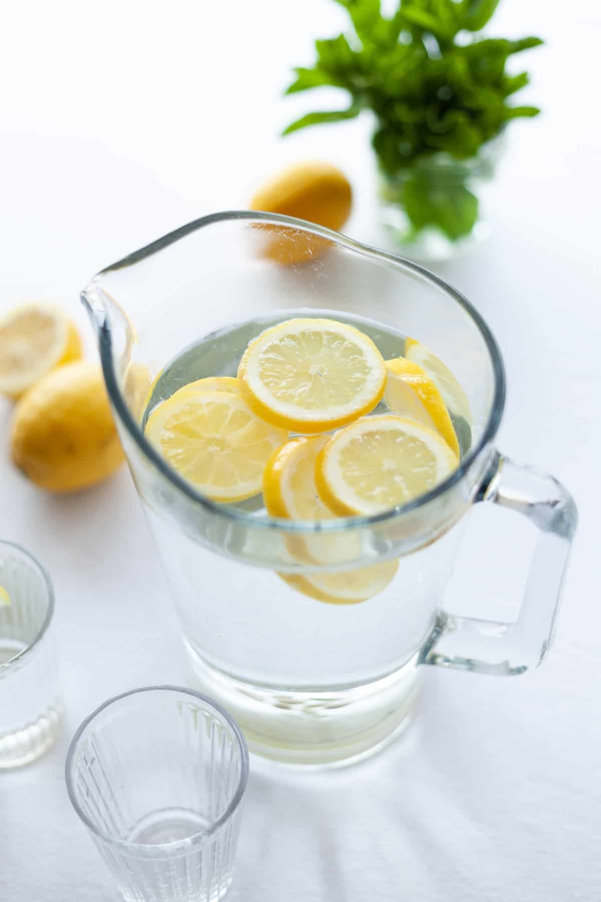 45 degree angle shot of clear glass pitcher filled with fruit water on a white table with halved lemons and a bunch of herbs in the background. photo credit julia zolotova.