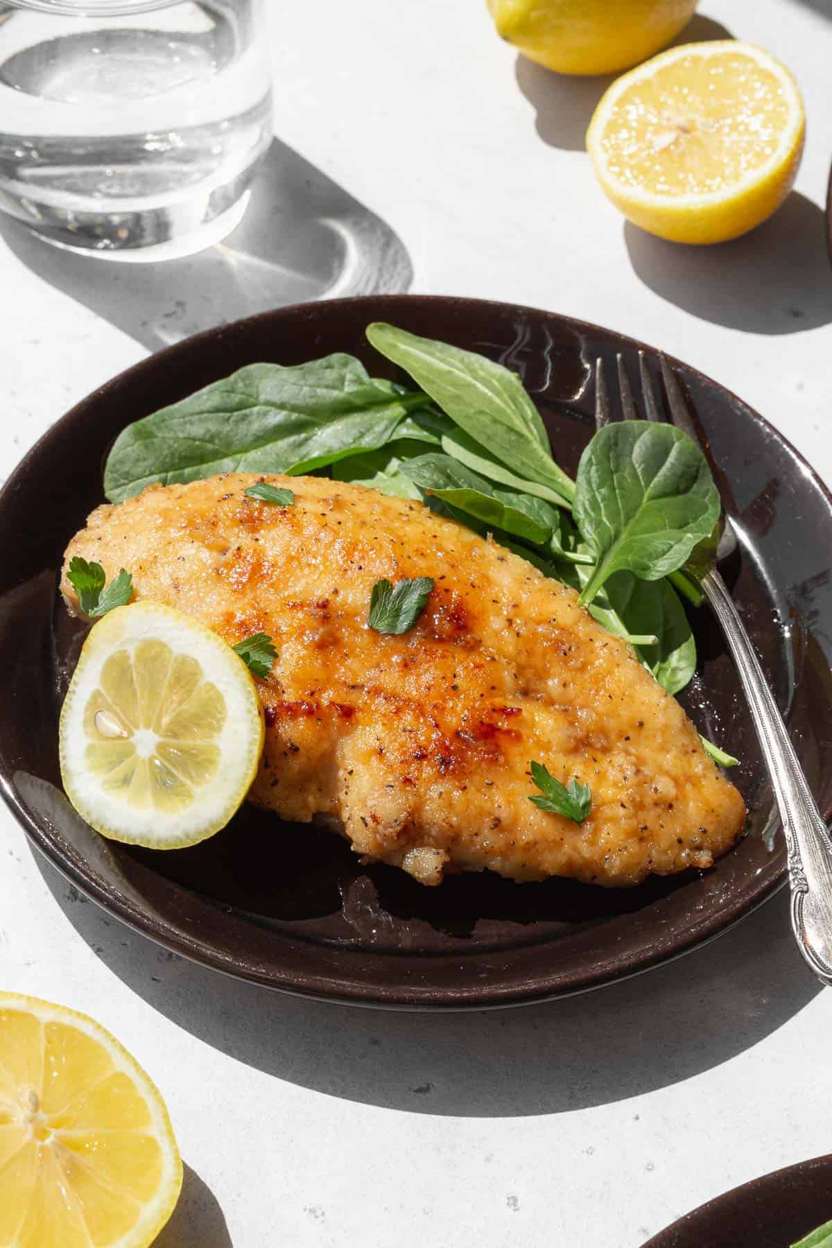 45 degree angle shot of a serving of lemony Italian chicken al limone on a black plate garnished with a slice of lemon and fresh parsley next to a simple spinach salad.