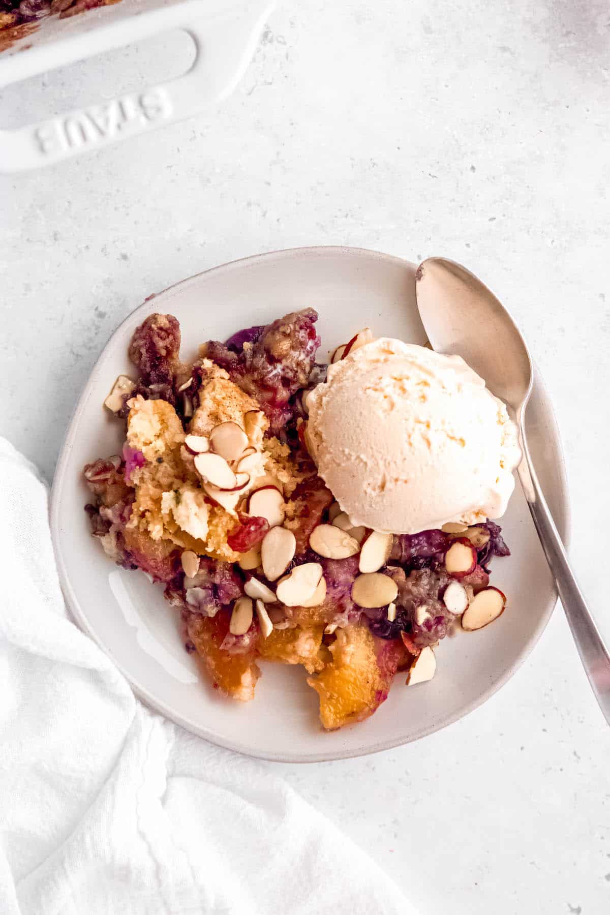 single serving of peach blueberry dump cake on a white dessert plate with a scoop of ice cream and a silver spoon.