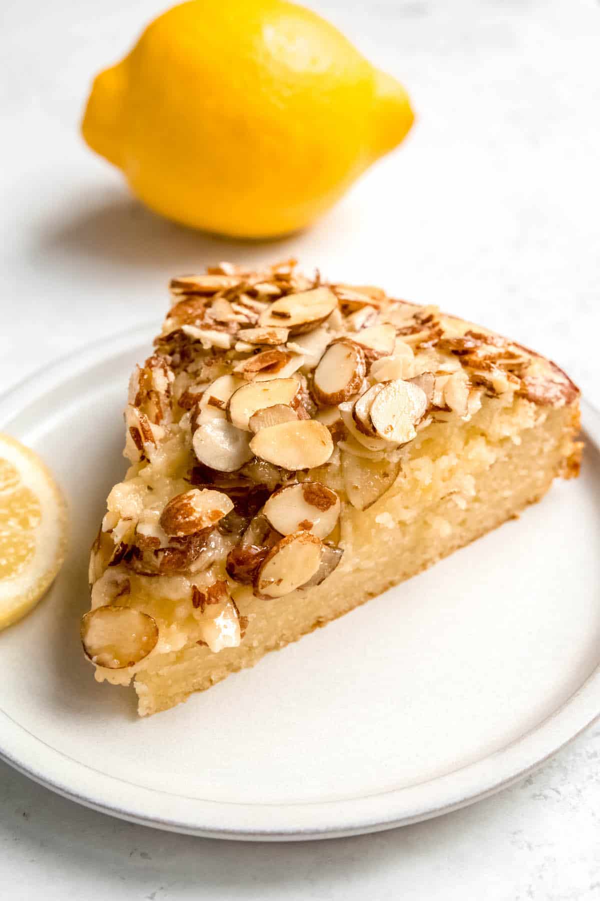 slice of Italian ricotta cake with lemons and almonds on a white dessert plate with a lemon in the background.