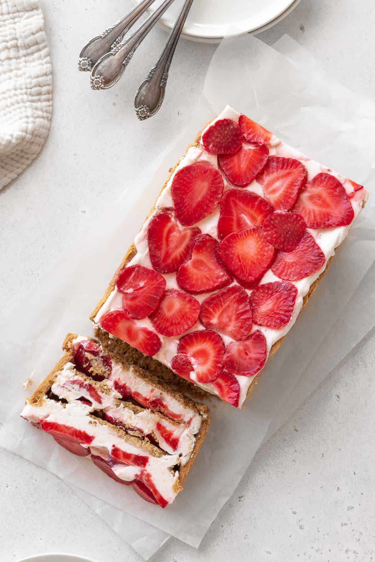 overhead shot of strawberry cream cheese icebox cake on a piece of parchment with one slice tipped over to show the layers of the no-bake strawberry graham cracker dessert.