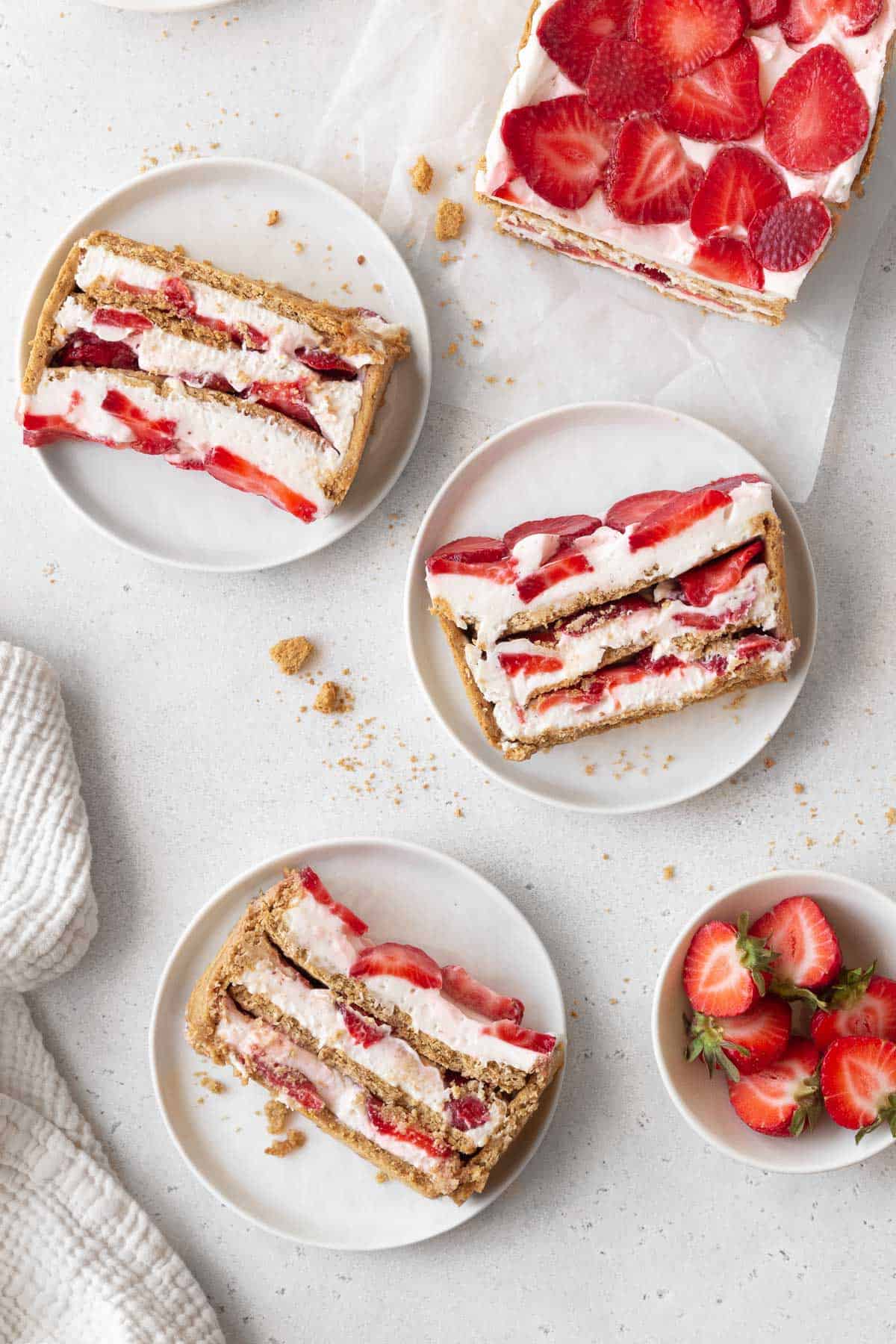 overhead shot of the strawberry cream cheese icebox cake on a piece of parchment with 3 slices on white dessert plates next to a bowl of halved strawberries.