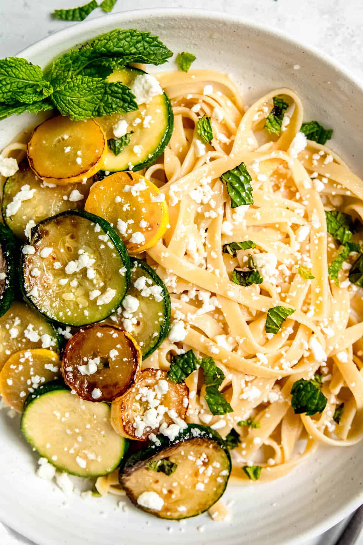 closeup hero shot of a bowl of zucchini linguini with rounds of cooked summer squash, tender pasta, crumbled feta, and chopped mint.