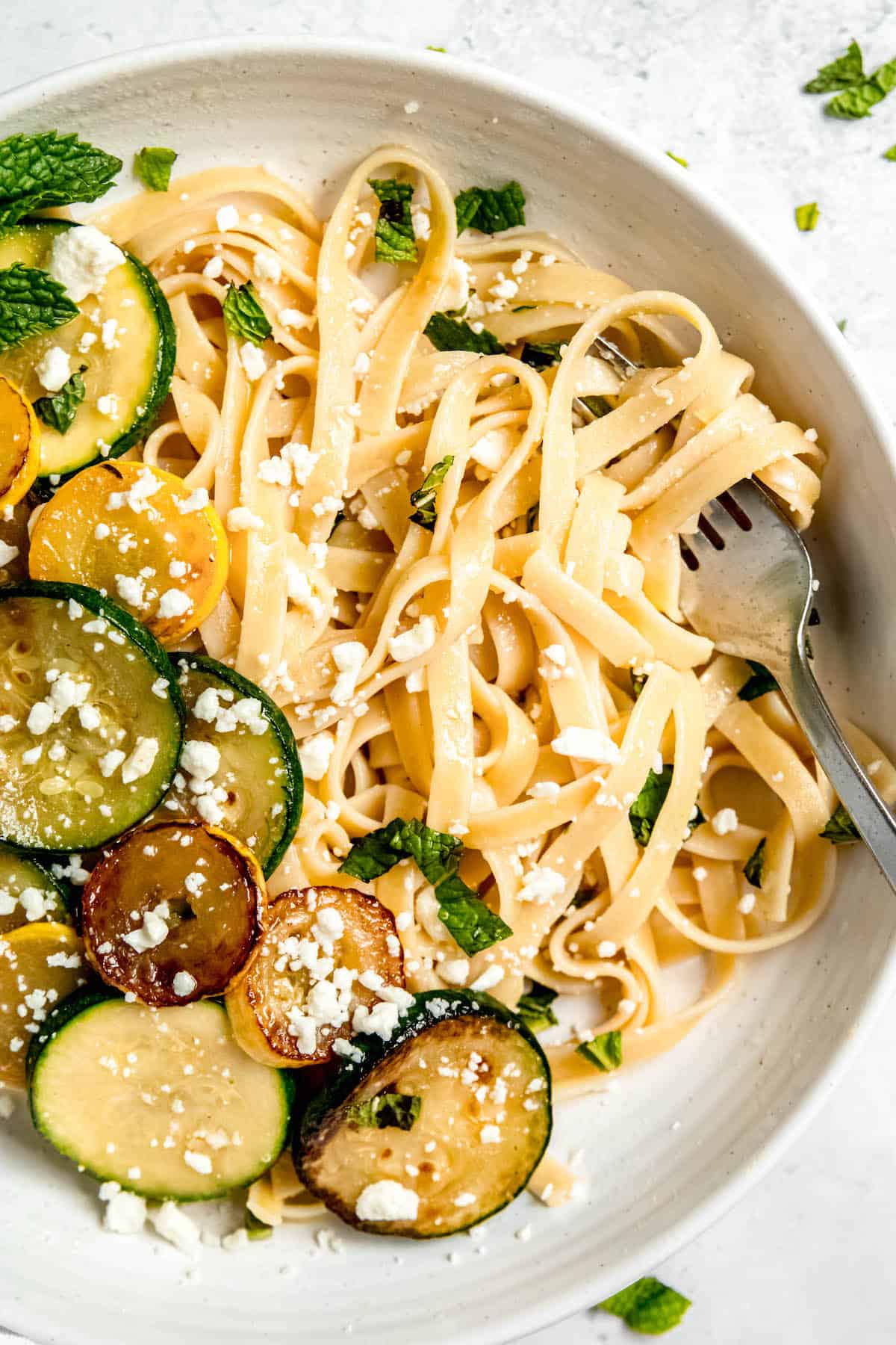 closeup hero image of a white pasta bowl with zucchini linguini with a silver fork twirled into the summer pasta.