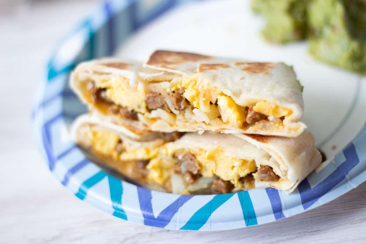 DIY frozen breakfast burritos sliced in half to show the filling and plated with guacamole on a paper plate.