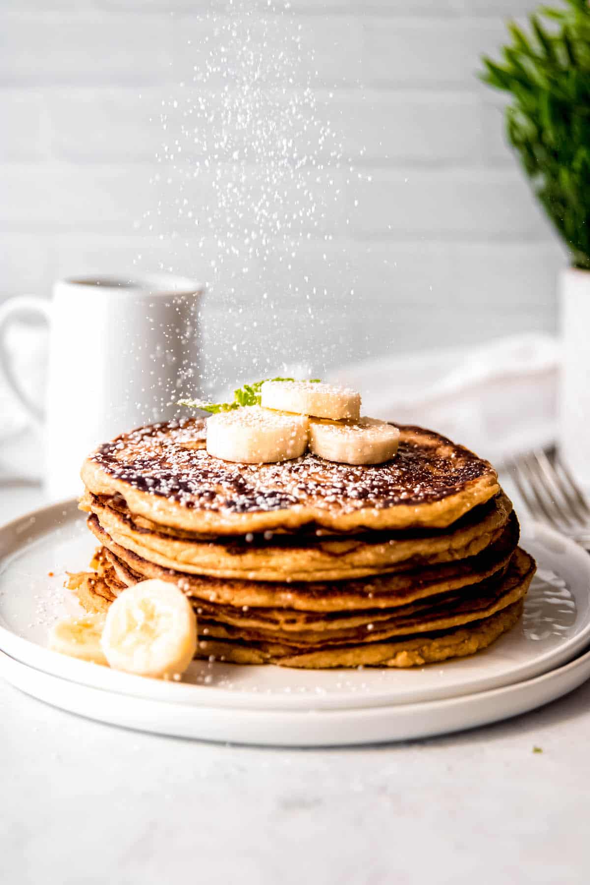 action shot of powdered sugar being sprinkled on a stack of oatmeal banana protein pancakes made with cottage cheese.
