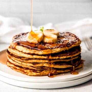 Square hero image of a stack of banana oatmeal protein pancakes being drizzled with maple syrup.