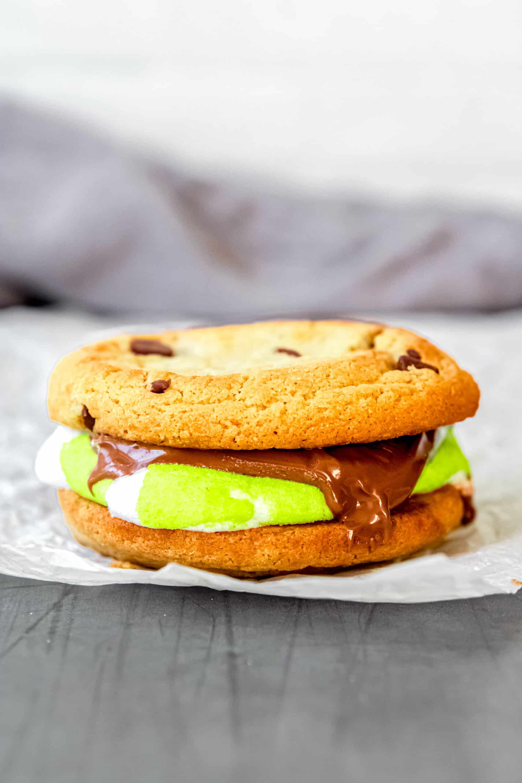 s'more made with a green halloween peep and melted chocolate peanut butter cup sandwiched between two chocolate chip cookies.