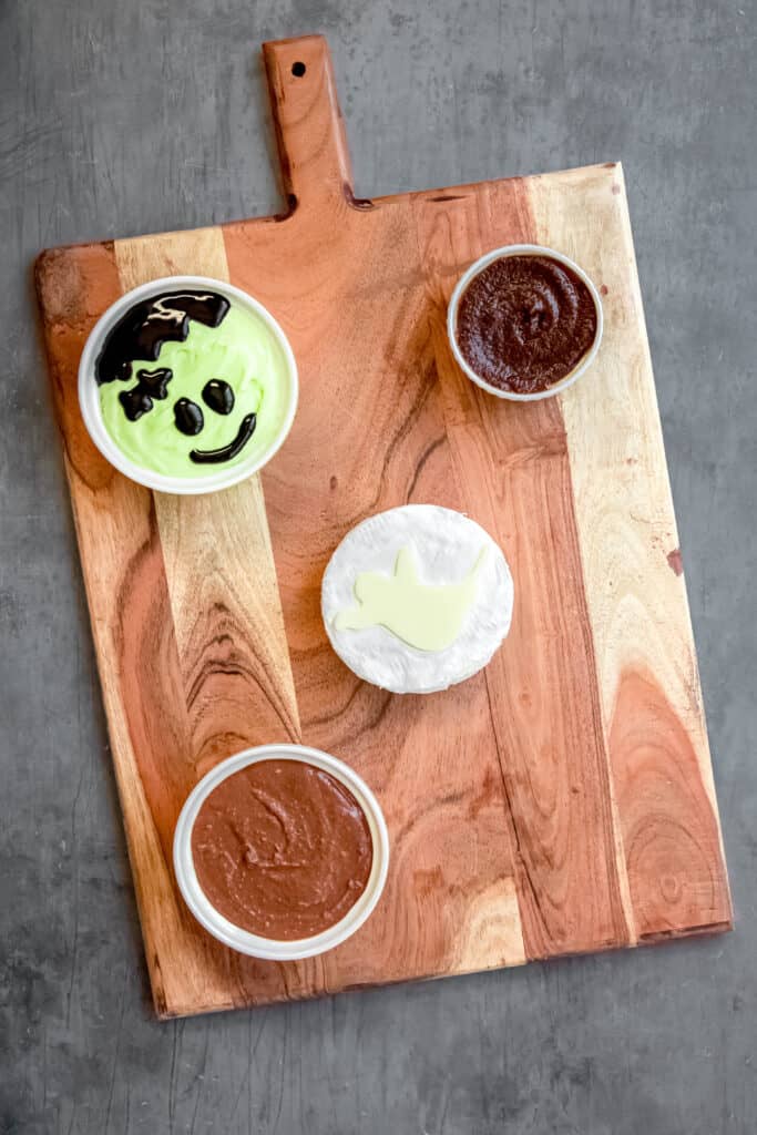 cream cheese fruit dip with a frankenstein face, round of brie with a ghost cutout on top, a bowl of chocolate hummus, and a bowl of apple butter placed as anchors on a large wooden charcuterie board to make a Halloween-themed s'mores board.