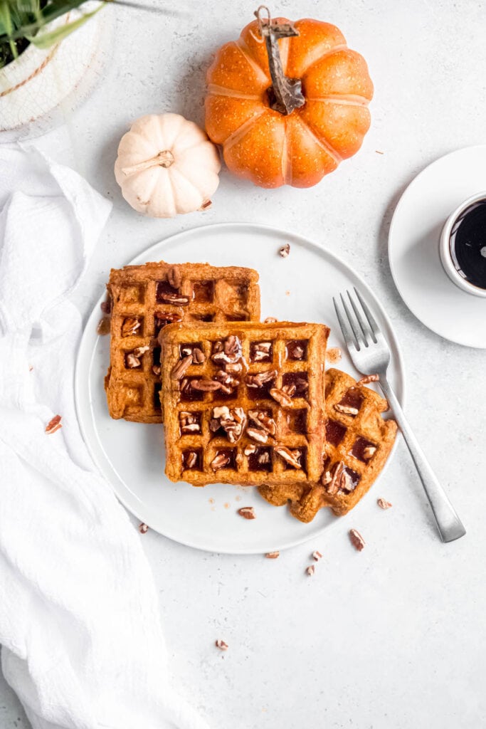 breakfast table set with two decorative pumpkins, a cup of coffee on a saucer, and 3 pumpkin pecan waffles on a white plate.