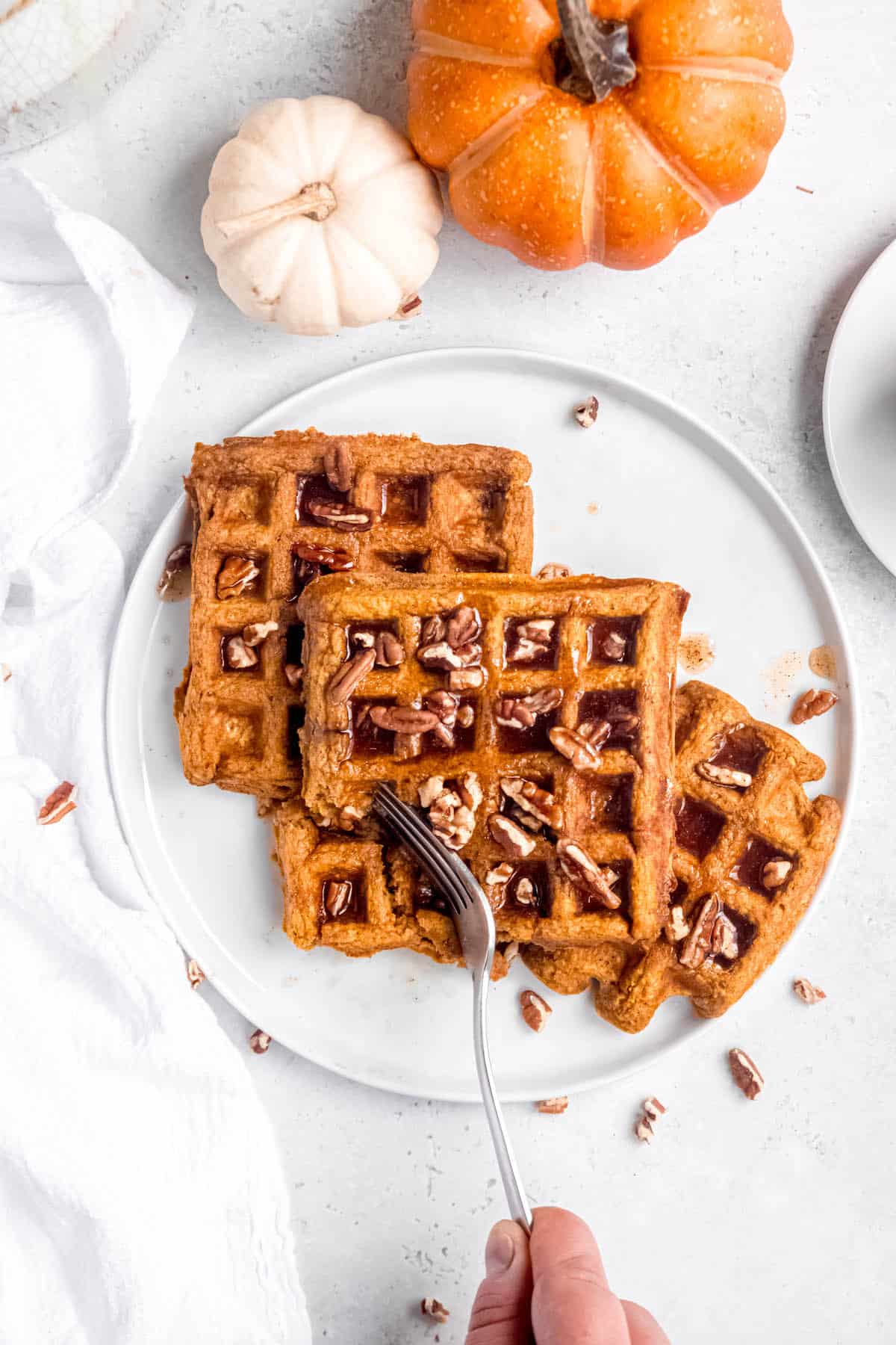 hero shot of a hand using a silver fork to cut a bite out of the plate of pumpkin pecan waffles.
