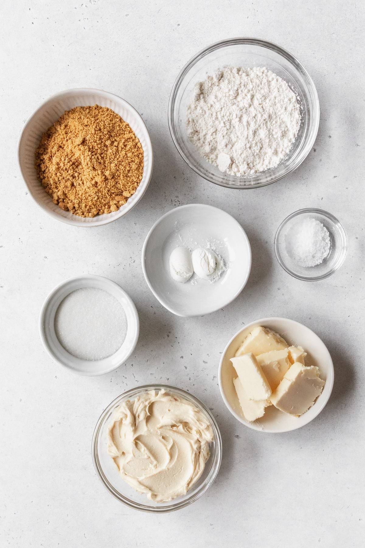 ingredients to make the graham cracker crumble and the cheesecake swirl measured out into bowls on a white table.