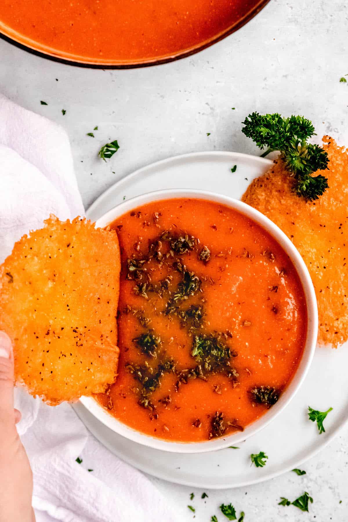hand dipping a gouda cheese crisp into a bowl of roasted tomato and red pepper soup.