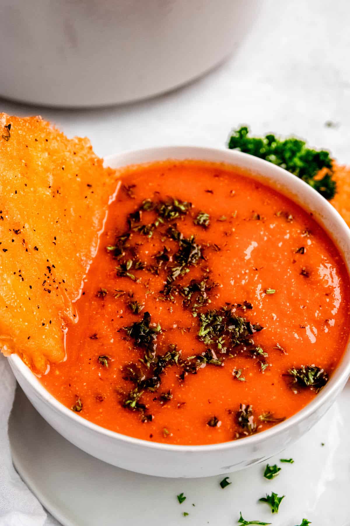 45 degree angle of a bowl of roasted red pepper soup with gouda cheese crisp dunked in the side and chopped parsley as a garnish.