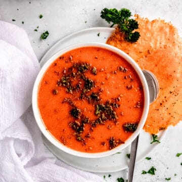 square hero image of roasted red pepper soup with gouda frico on a white table.