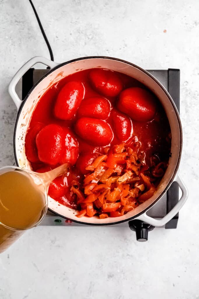 whole canned tomatoes and their juices, drained red peppers, and chicken bone broth being added to the dutch oven with the sautéed aromatics.