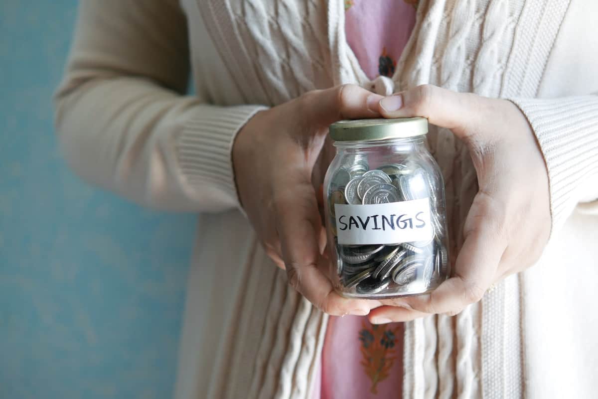 hands holding a small mason jar with coins that has a sticker that reads "Savings" on the front.