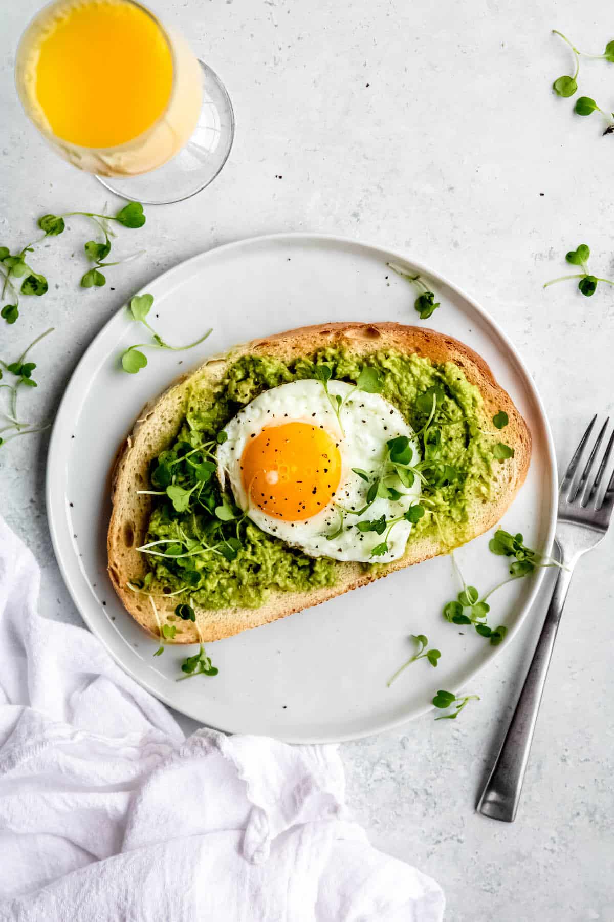 flat lay hero shot of an avocado tartine topped with a sunny side up egg on a white plate garnished with microgreens next to an orange mimosa.