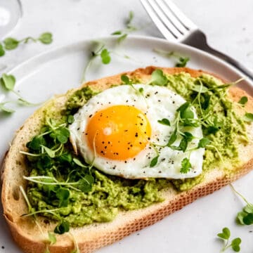45 degree angle hero image of a fried egg and avocado tartine on a white plate with microgreens and a silver fork.