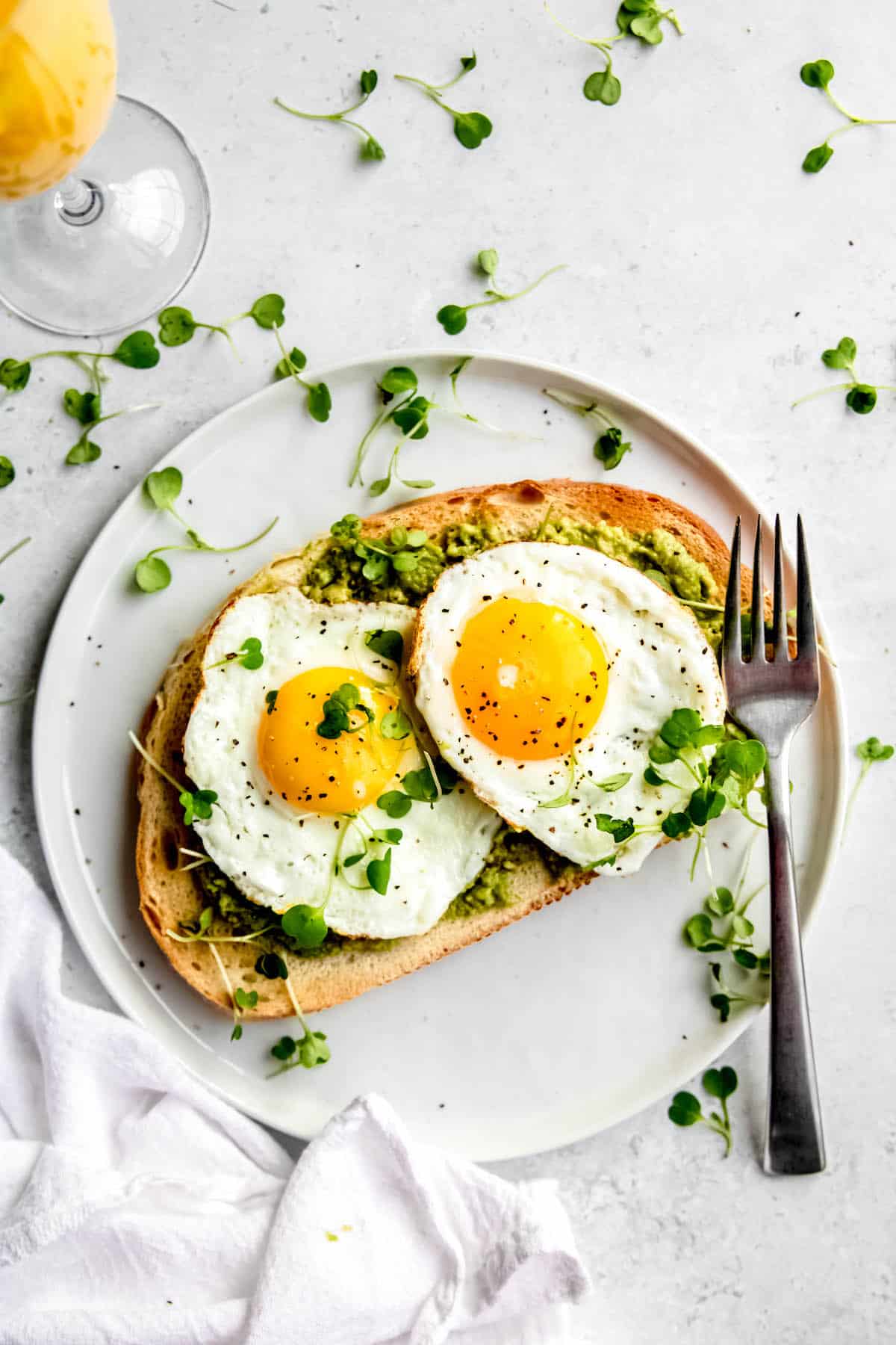 flat lay shot of an avocado tartine with 2 fried eggs next to an orange mimosa on a white table with microgreens scattered around.