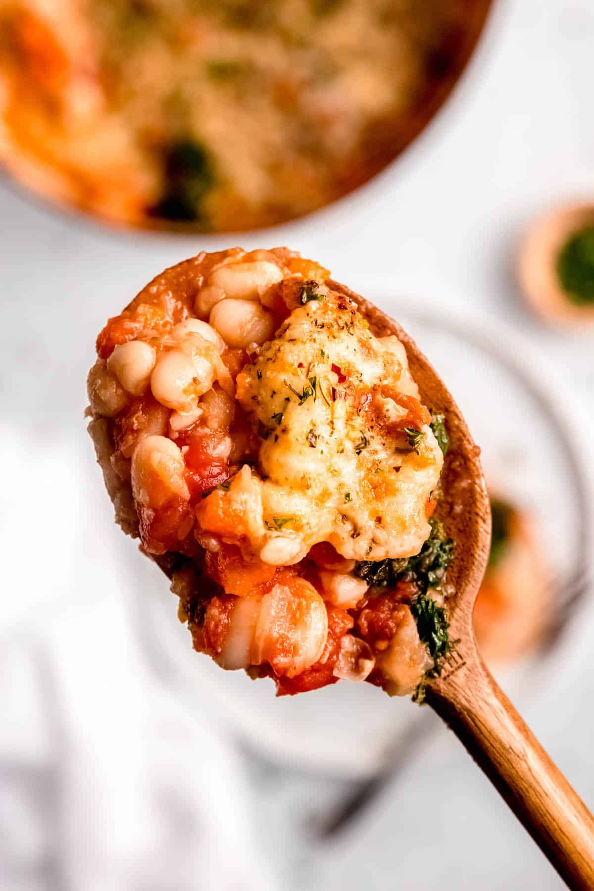 closeup shot of a wooden spoon with a scoop of the cheesy tomato bean bake showing the browned cheese, saucy tomato beans, and bright kale.