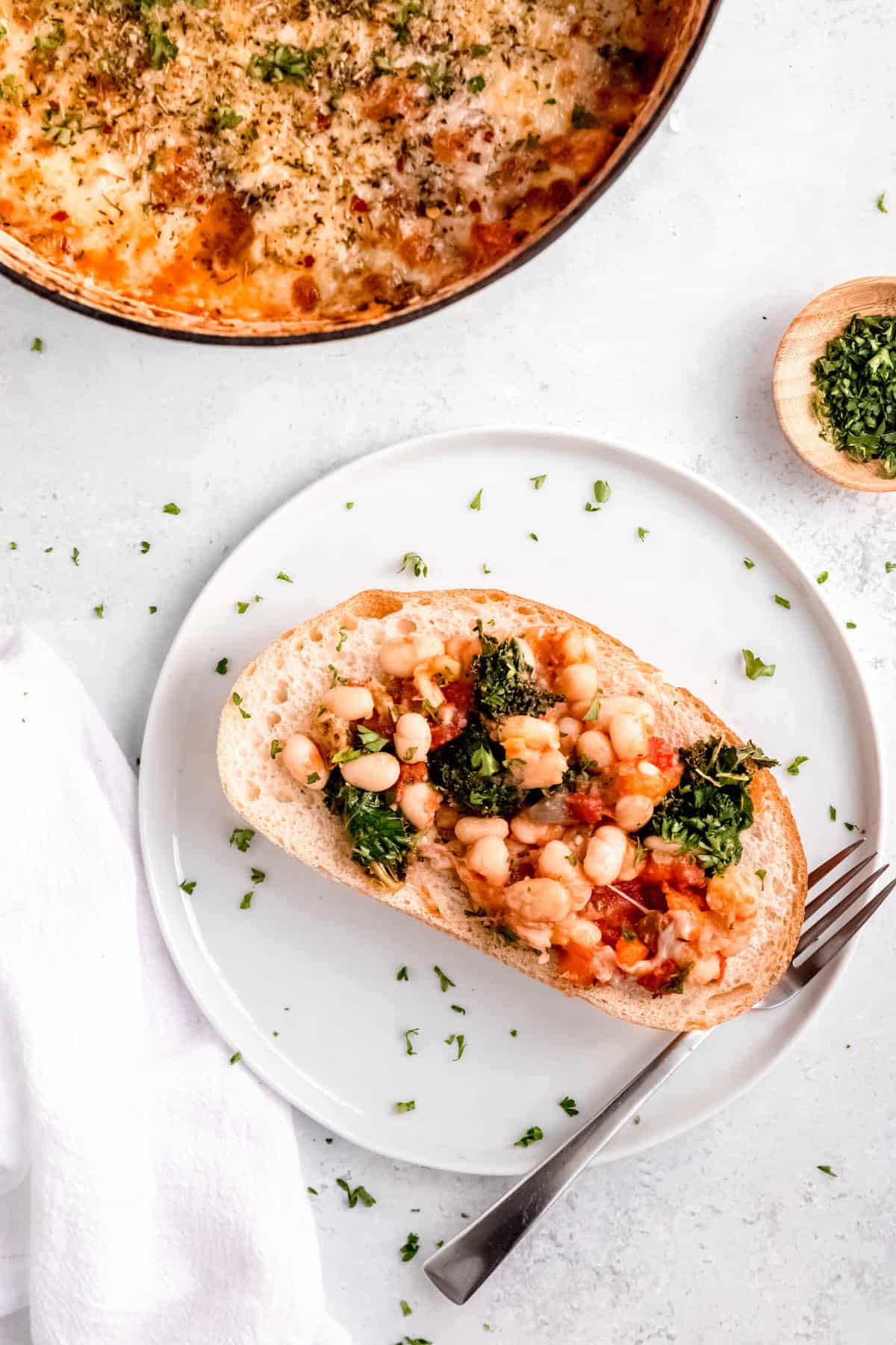 flat lay shot of a serving of cheesy pizza beans served on a piece of crusty toast with the rest of the cheesy white bean tomato bake in a dutch oven to the side.