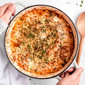 square hero image of two hands grabbing a Dutch oven filled with pizza-flavored cheesy white bean tomato bake made with dried beans.