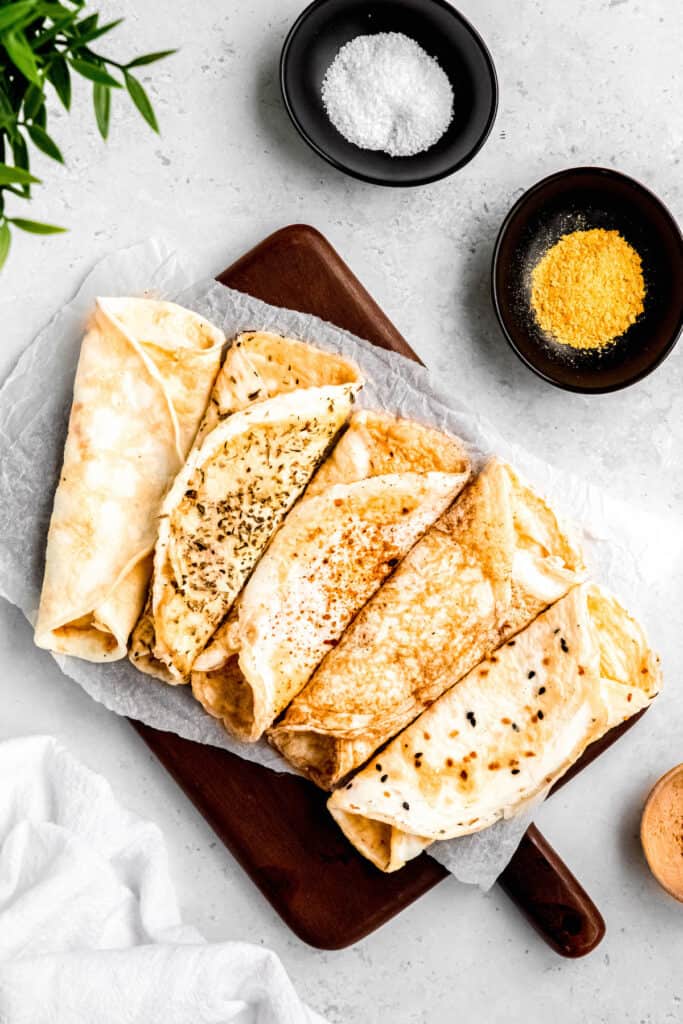 flat lay shot of egglife copycat egg white wraps in all five flavors, fanned out on a wooden cutting board with small bowls of kosher salt and nutritional yeast.