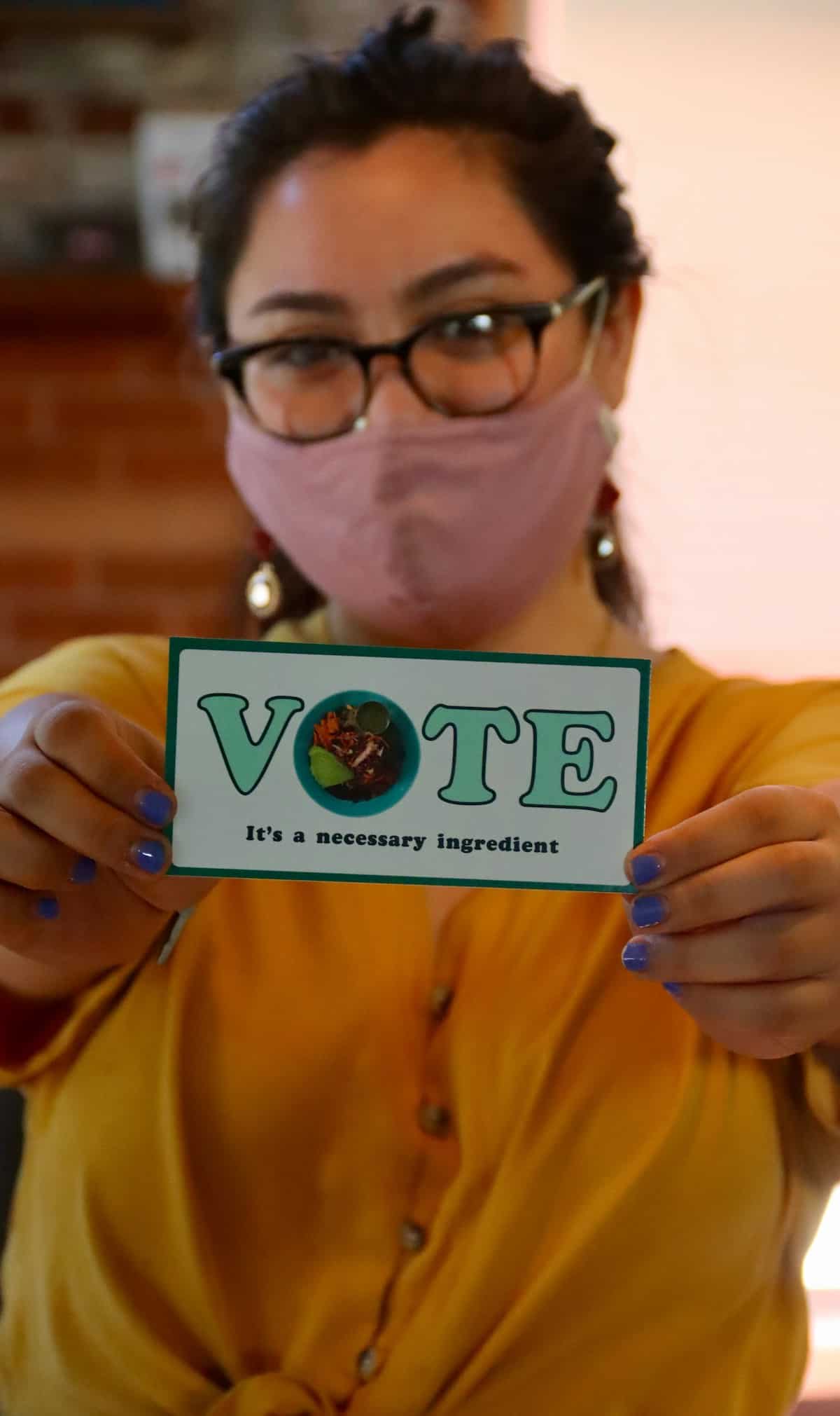 Woman holding a card that says "Vote: It's a necessary ingredient".