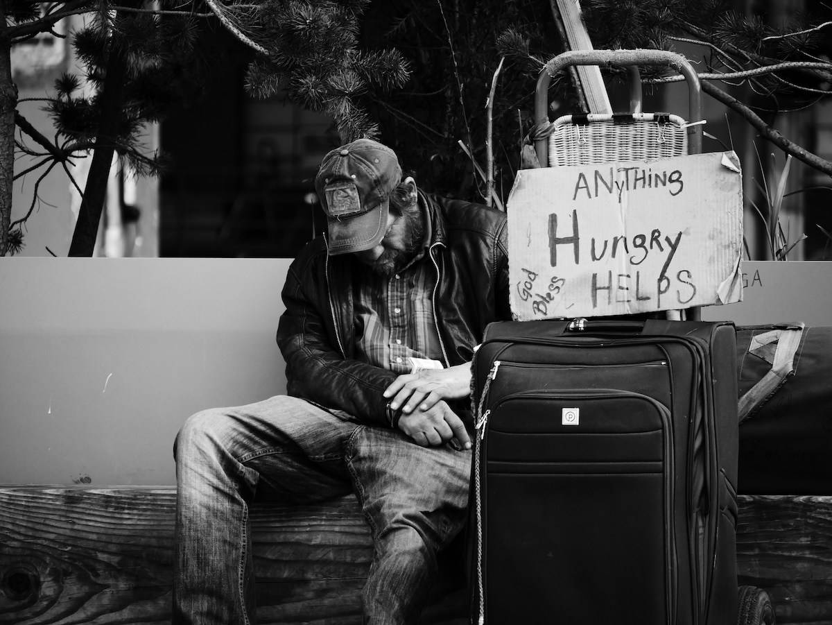homeless man on a bench with a sign that says "Hungry, anything helps."