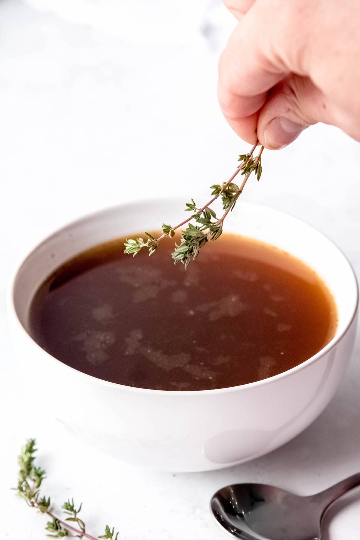hand adding a sprig of fresh thyme to a bowl of roasted bone broth for serving like consomme.