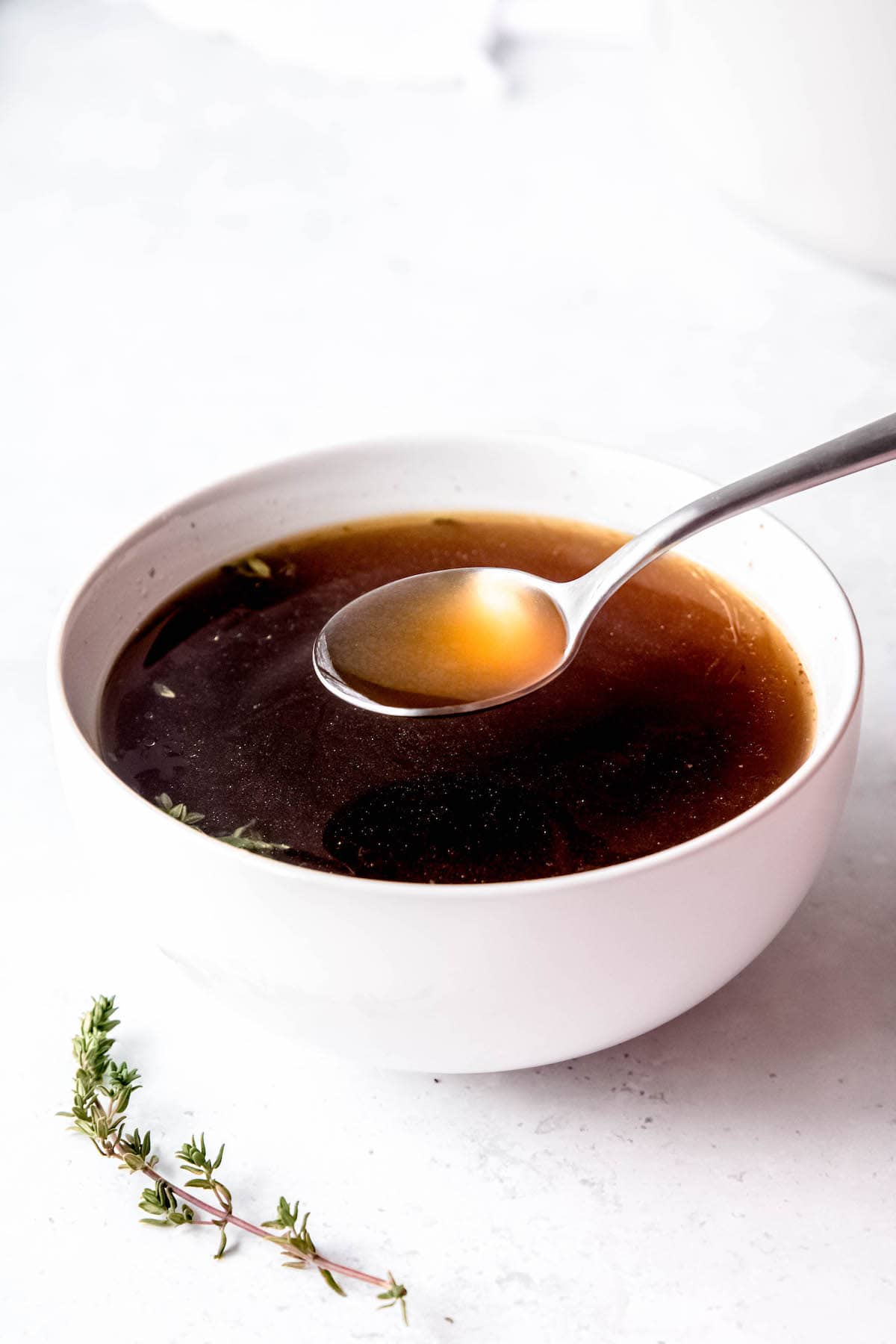 small white bowl filled with rich, dark beef brown bone broth from scraps with a soup spoon taking a bite.
