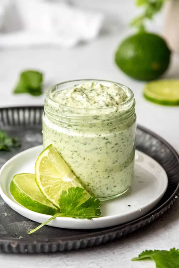 lime cilantro garlic sauce in a jar on a white plate on top of a pounded black round metal serving tray.