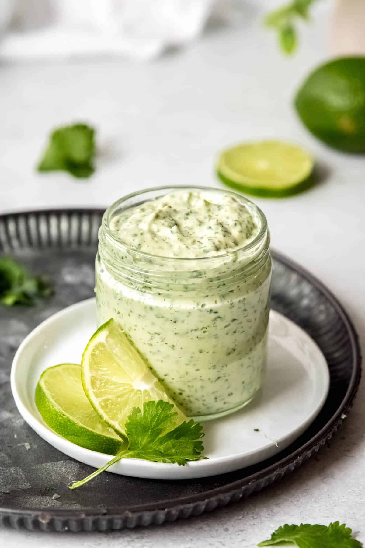 hero shot of a jar of pollo tropical style cilantro lime sauce with jalapenos and garlic on a white table with slices of limes and fresh cilantro strewn about.