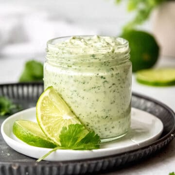 square hero image of a glass jar of creamy lime garlic cilantro sauce on a white plate with lime slices and a sprig of fresh cilantro.