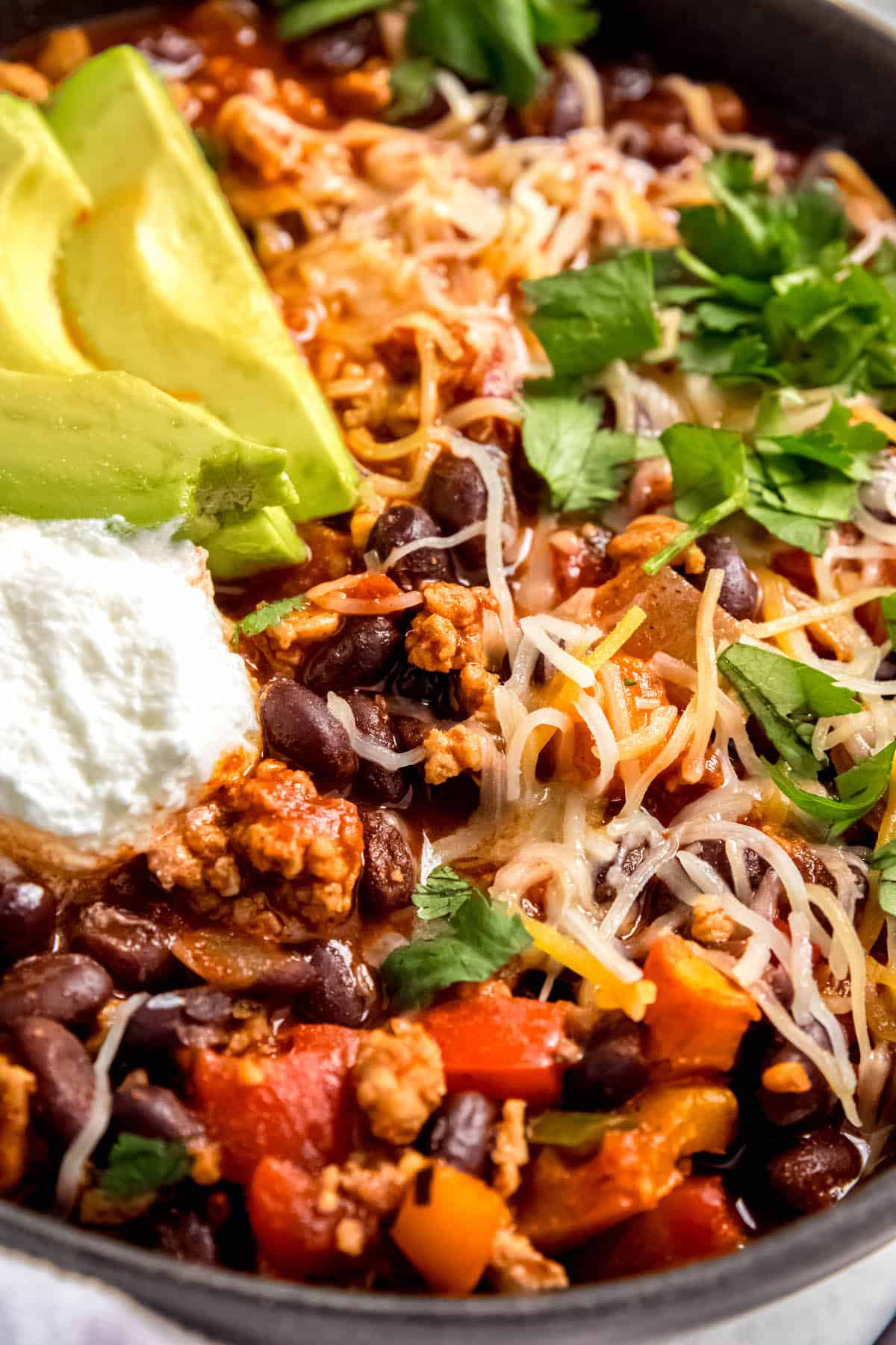 45 degree angle of a bowl of garnished black bean chicken chili topped with shredded cheddar and avocado.