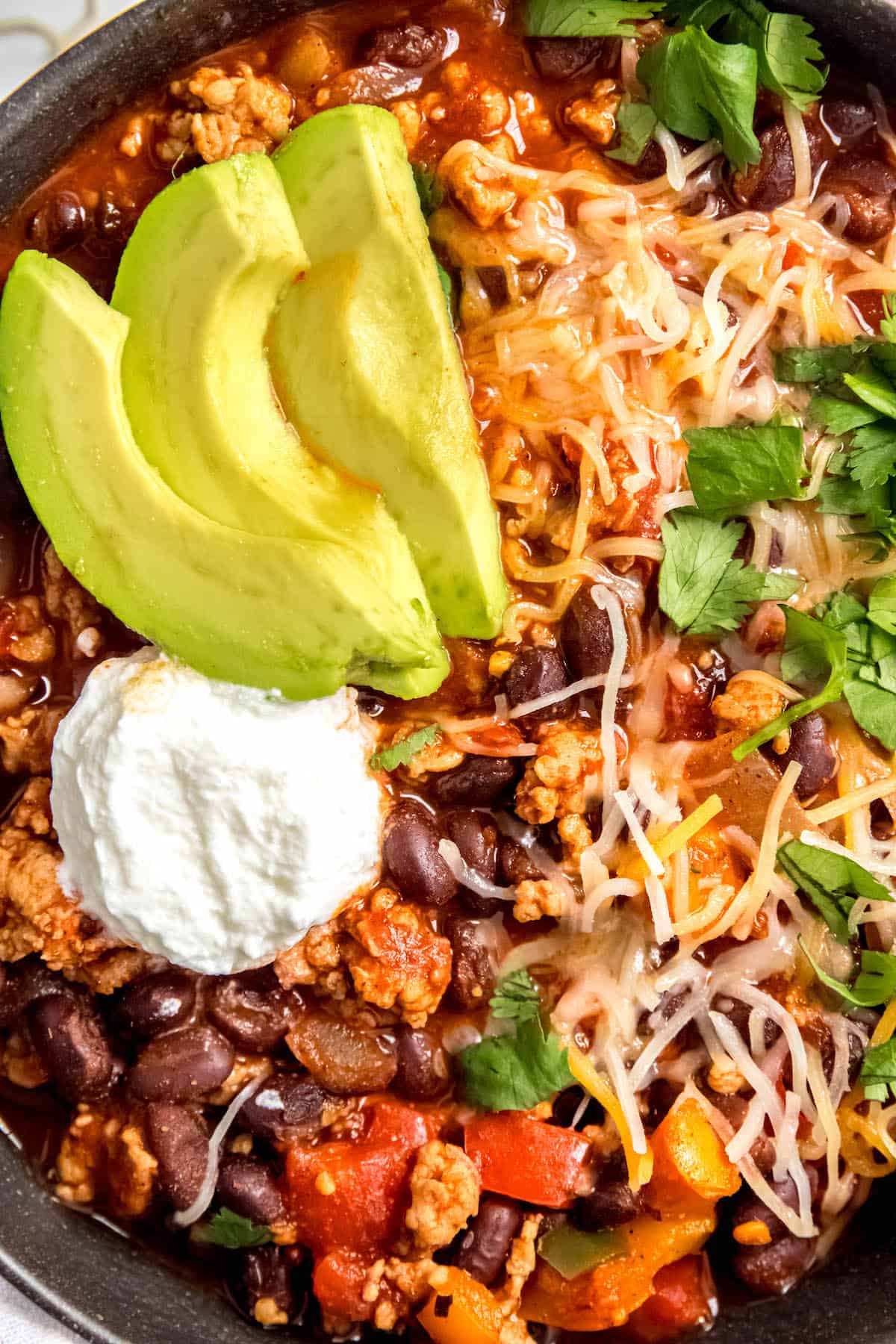 closeup overhead shot of spicy chicken and black bean chili topped with sliced avocado, greek yogurt, and shredded cheese.