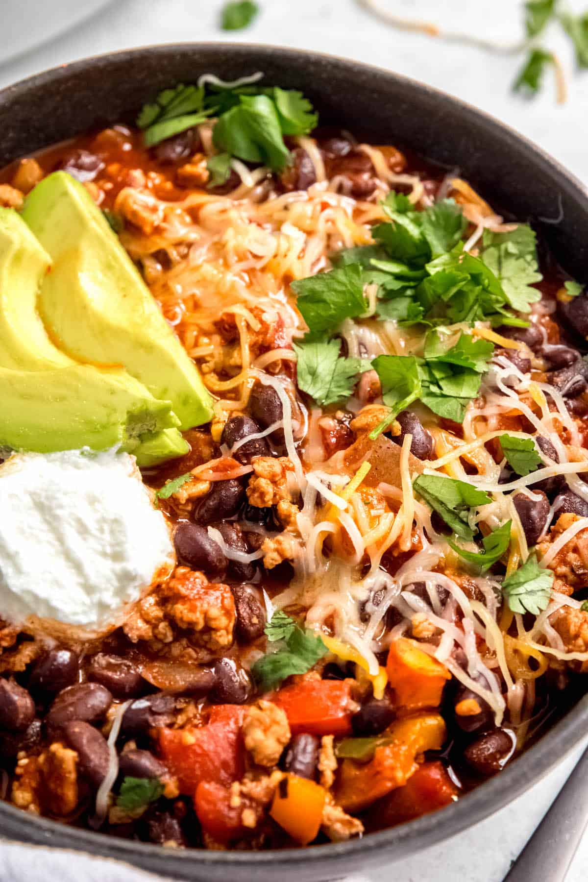 closeup shot of a bowl of chicken black bean chili with a dollop of sour cream, some sliced avocados, grated cheese, and chopped cilantro.