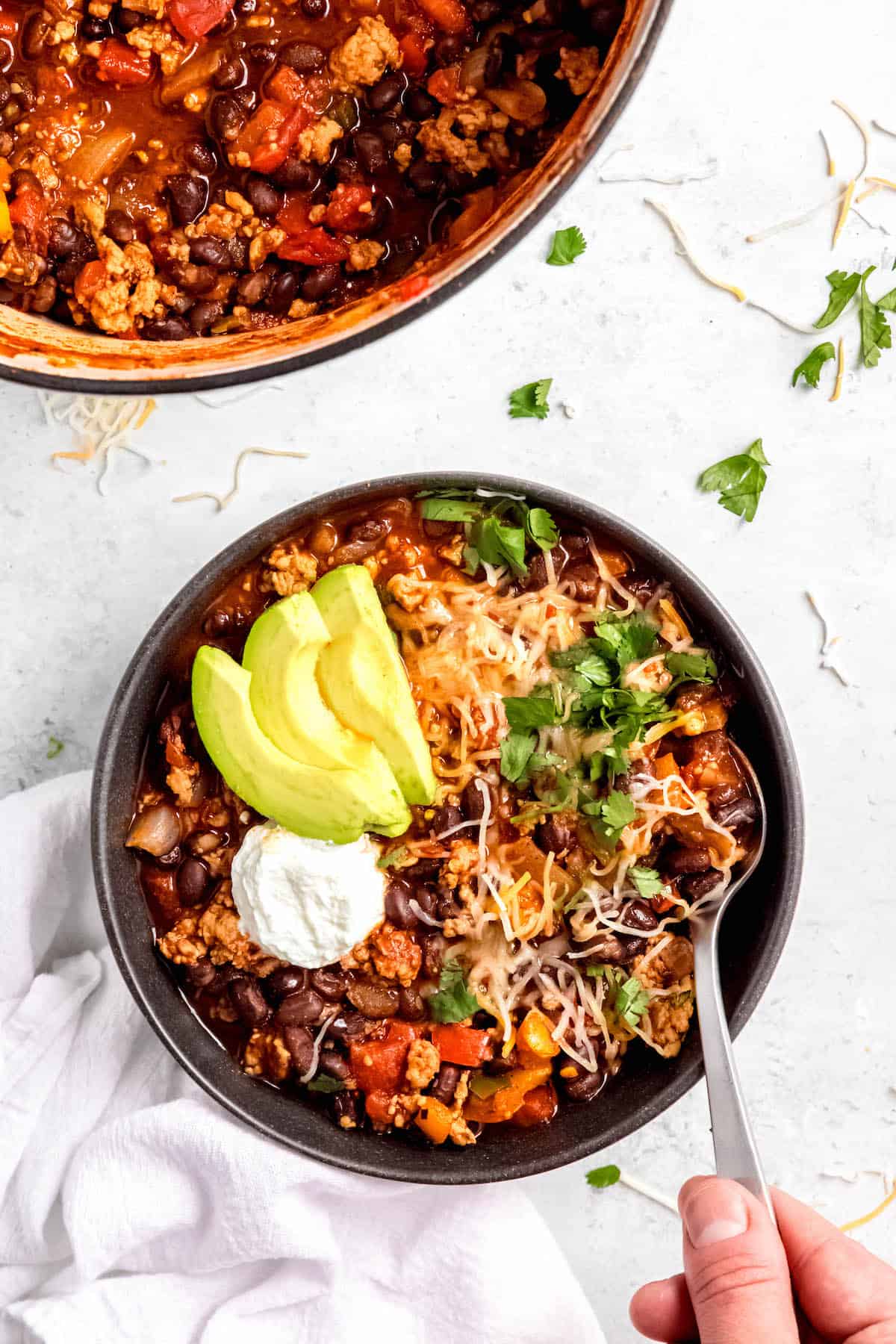 hero image of a black bowl of spicy black bean chicken chili with cheese, avocado, and cilantro garnishes with a hand holding a silver spoon.