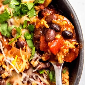 flat lay shot of a silver spoon taking a spoonful of chicken and black bean chili in a black bowl.