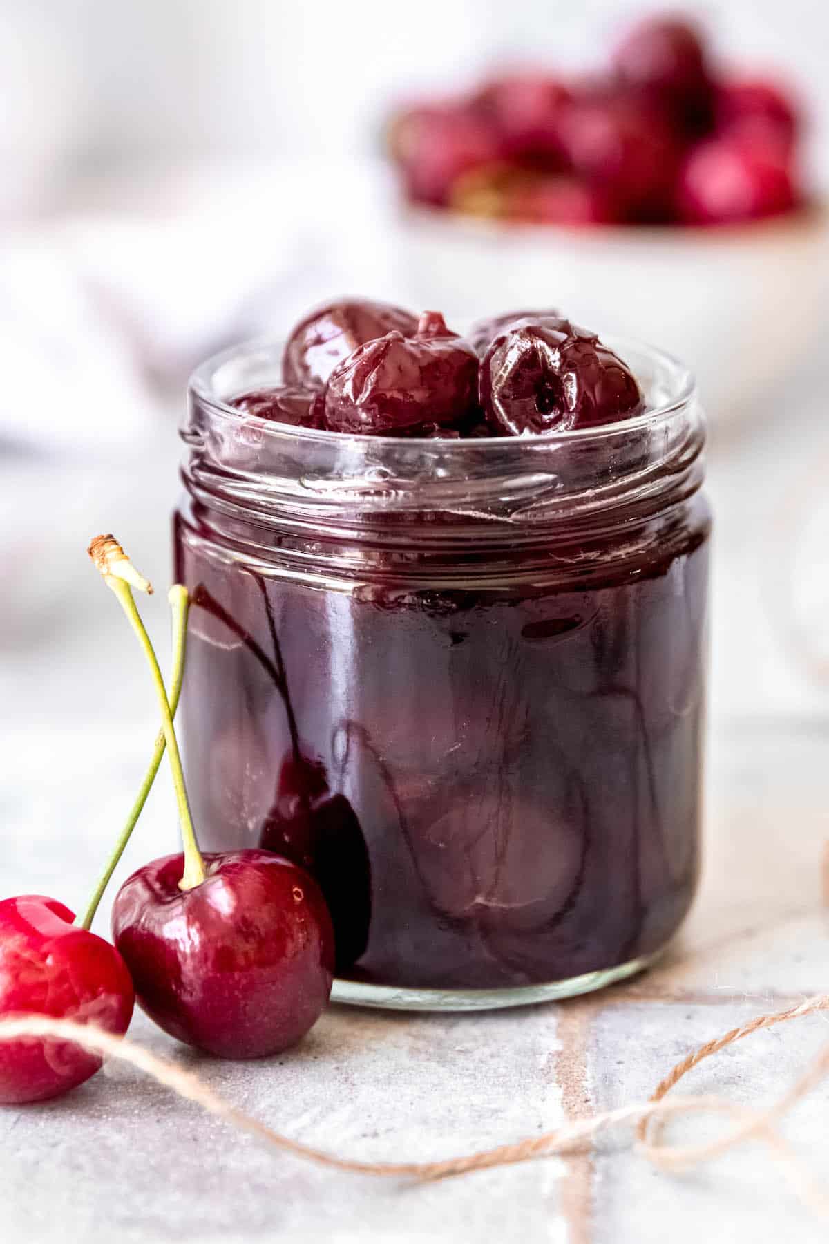 clear glass jar of homemade amaretto marinated cherries on a tiled surface with some twine and fresh cherries scattered around.
