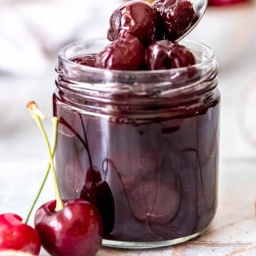 silver spoon taking a few amaretto cherries from a glass jar with fresh cherries in the foreground and background.
