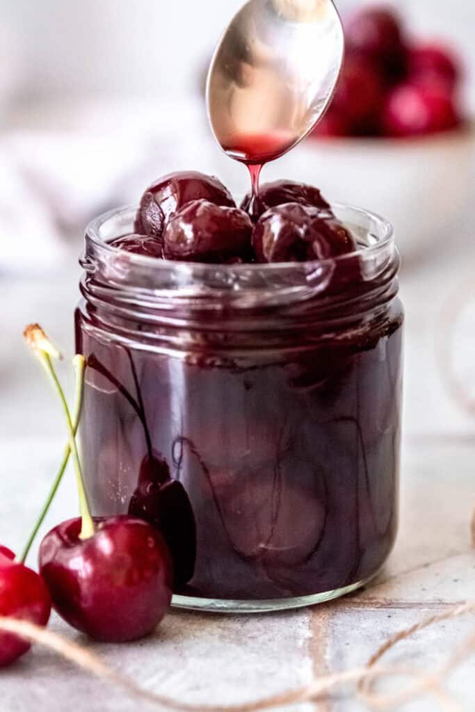 silver spoon drizzling the almond cherry syrup into a jar packed with homemade amaretto cherries.