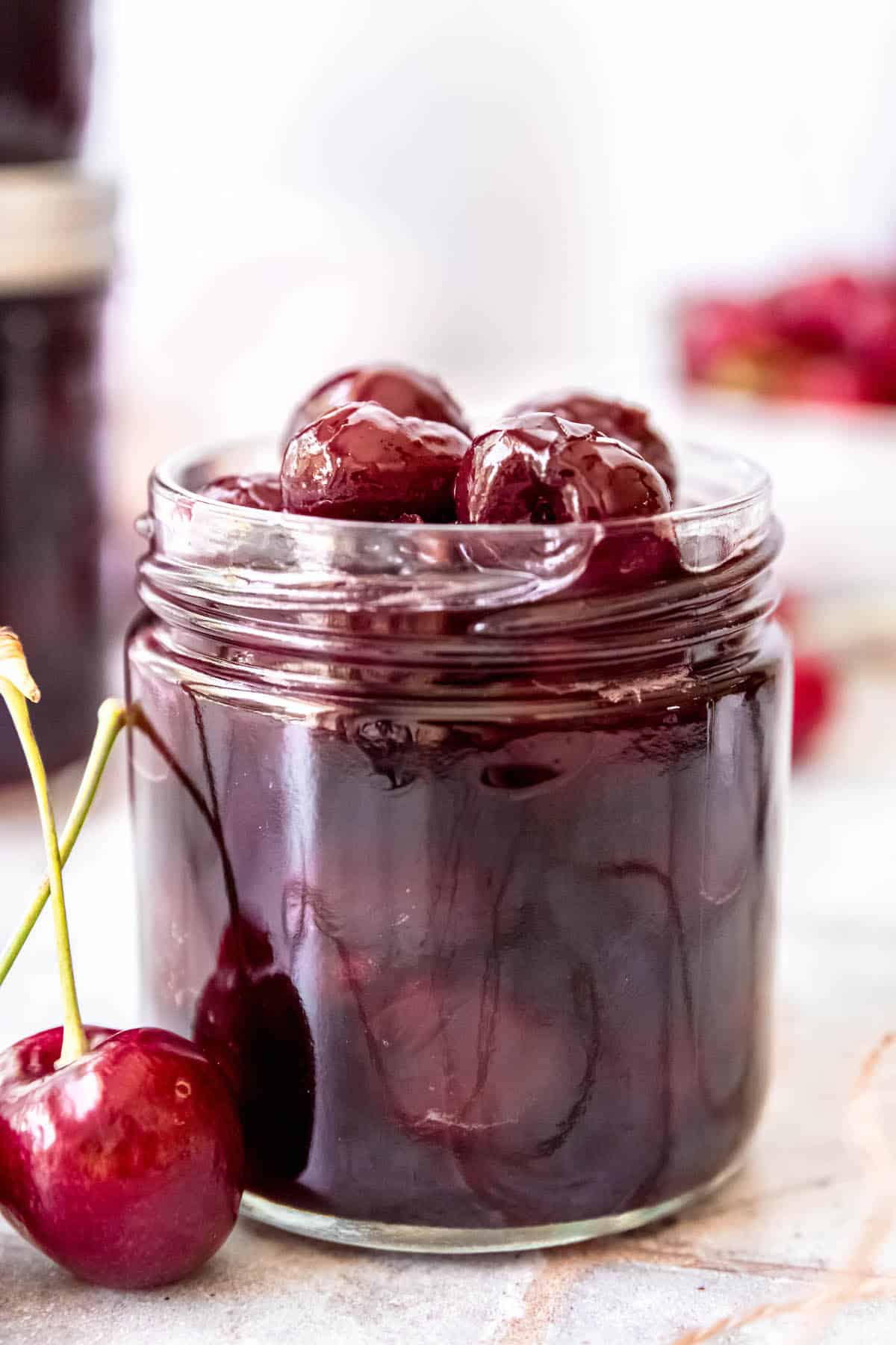 open glass jar of homemade amaretto cherries with a few jars of them stacked in the background and a bowl of fresh cherries blurred out.