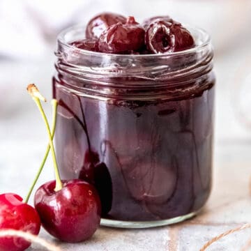 square hero image of homemade Lazzaroni amaretto cherries in a glass jar with 2 fresh cherries in front of it.