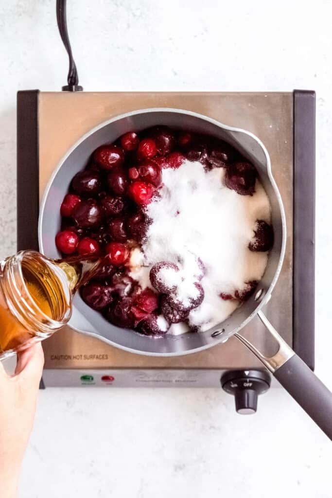 hand pouring amaretto liqueur into the saucepan with the cherries and sugar.